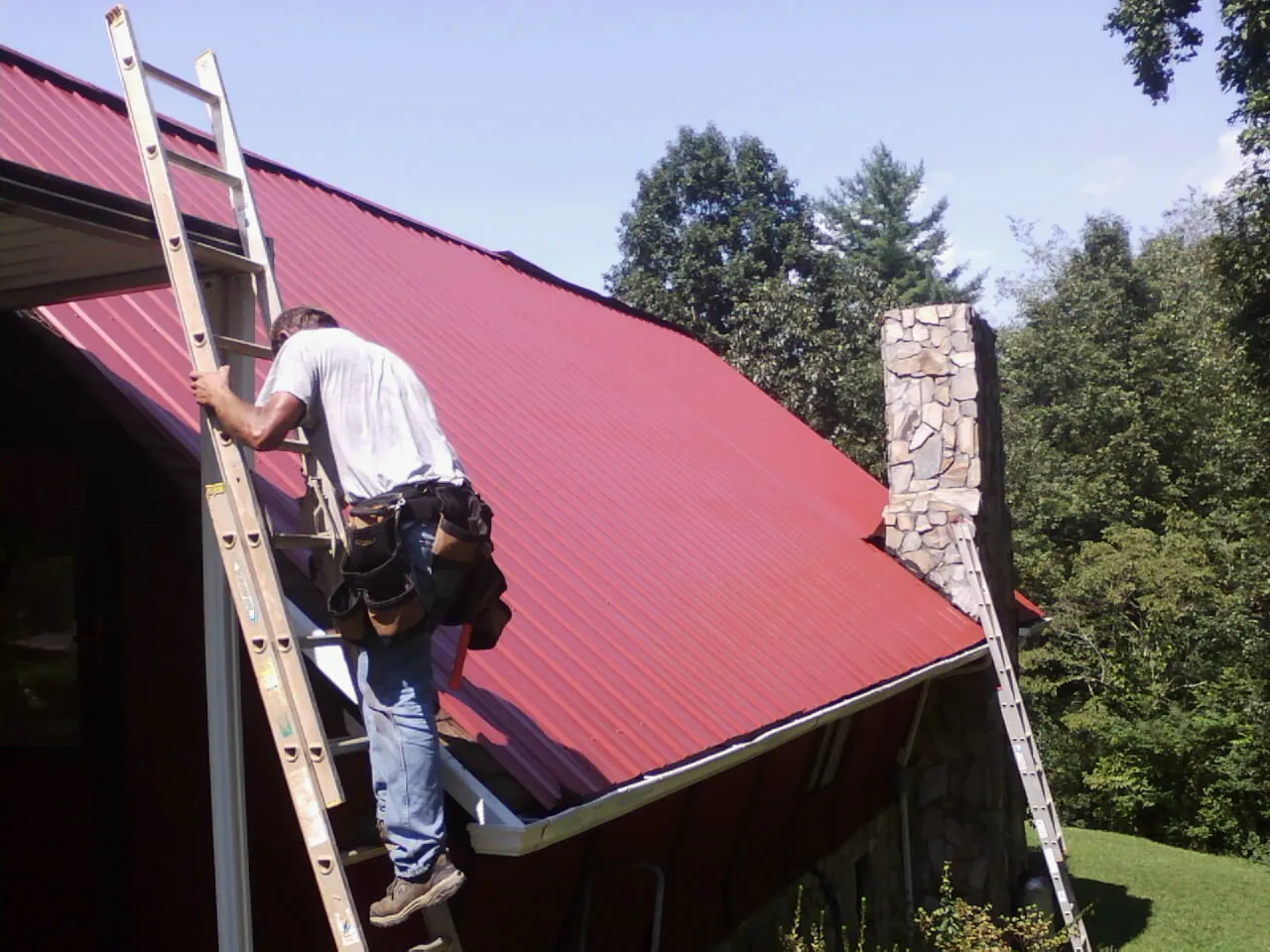 Roofer on a ladder beside a red metal roof and stone chimney — Soffit & Fascia Repair by Roofing Brothers in Wentworth
